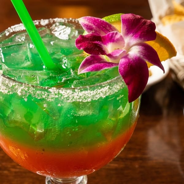A vibrant green and orange layered drink with a salted rim, garnished with a purple orchid and lime.