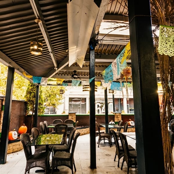 Outdoor patio dining area with wicker chairs, black tables, and colorful papel picado decorations.