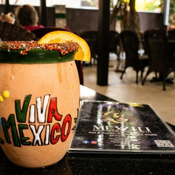 A "Viva Mexico" clay mug with a drink, orange slice, and chili rim, next to a menu.