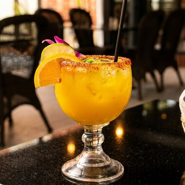 A vibrant yellow cocktail with a chili-lime rim, garnished with orange and lime slices, next to a basket of tortilla chips.