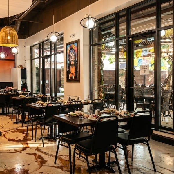 Restaurant interior with set tables, wicker pendant lights, and large windows looking out to a patio.