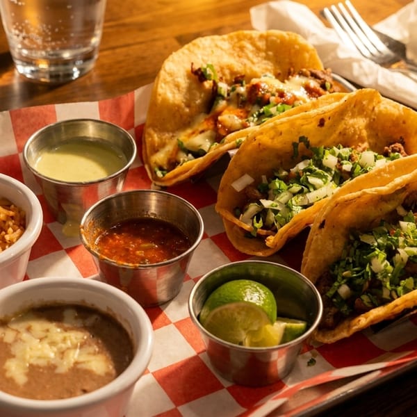 Three tacos with sides of rice, beans, salsa, and lime wedges on a checkered tray.