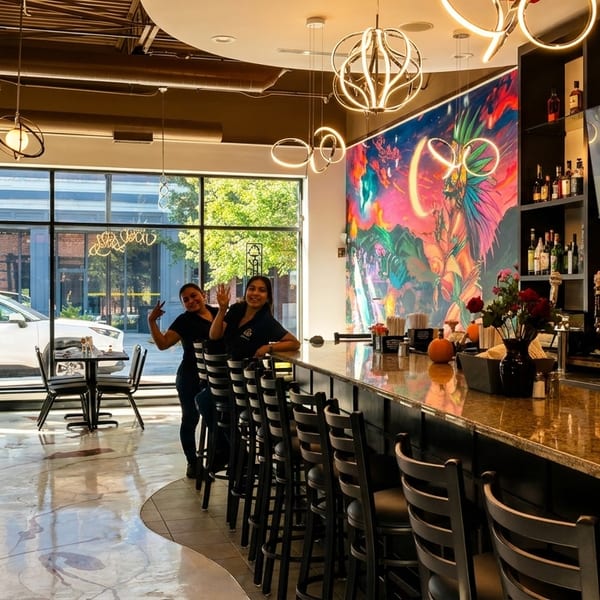 Two smiling women stand behind a long bar with stools, a colorful mural, and large windows.