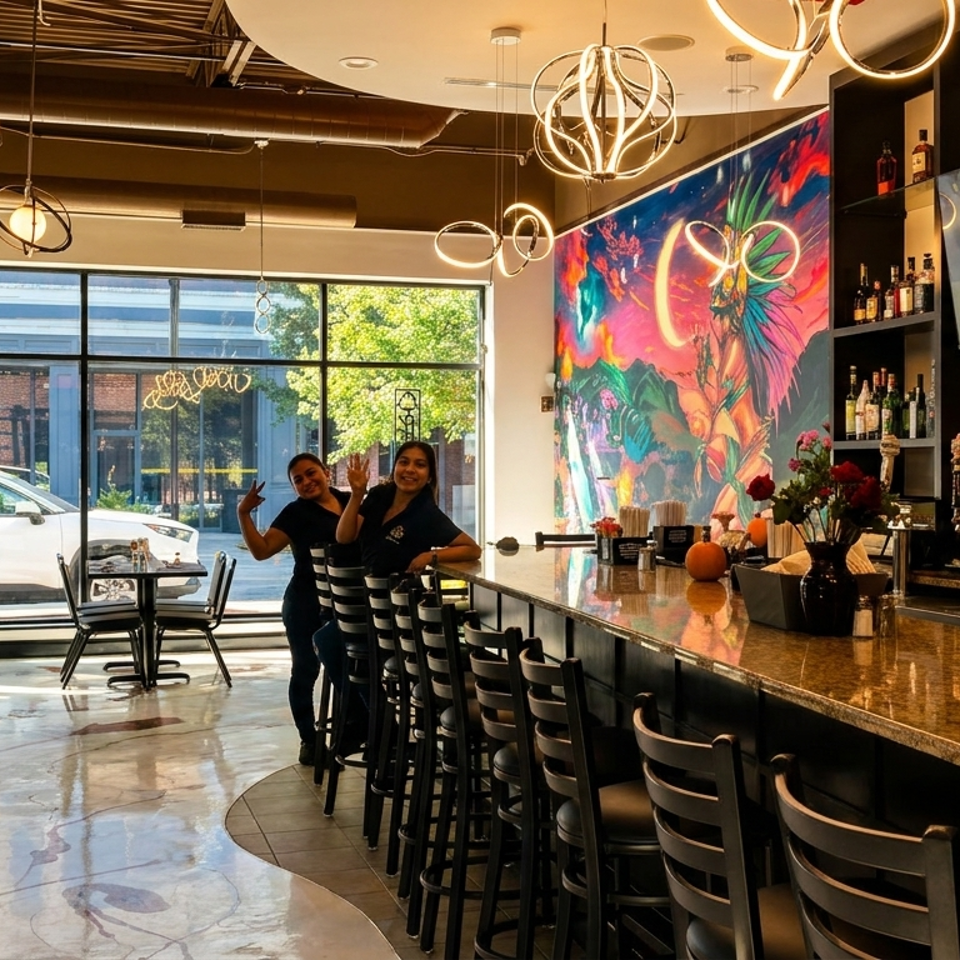 Two smiling women stand behind a long bar with stools, a colorful mural, and large windows.