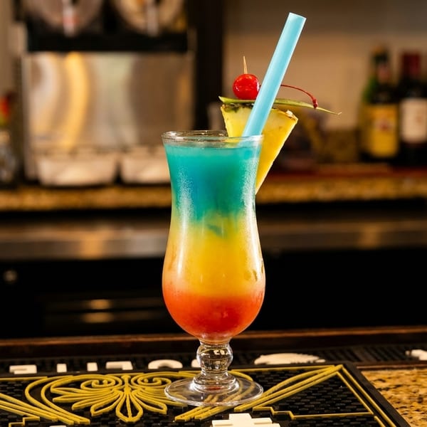 A colorful layered cocktail with pineapple and cherry garnish sits on a bar.